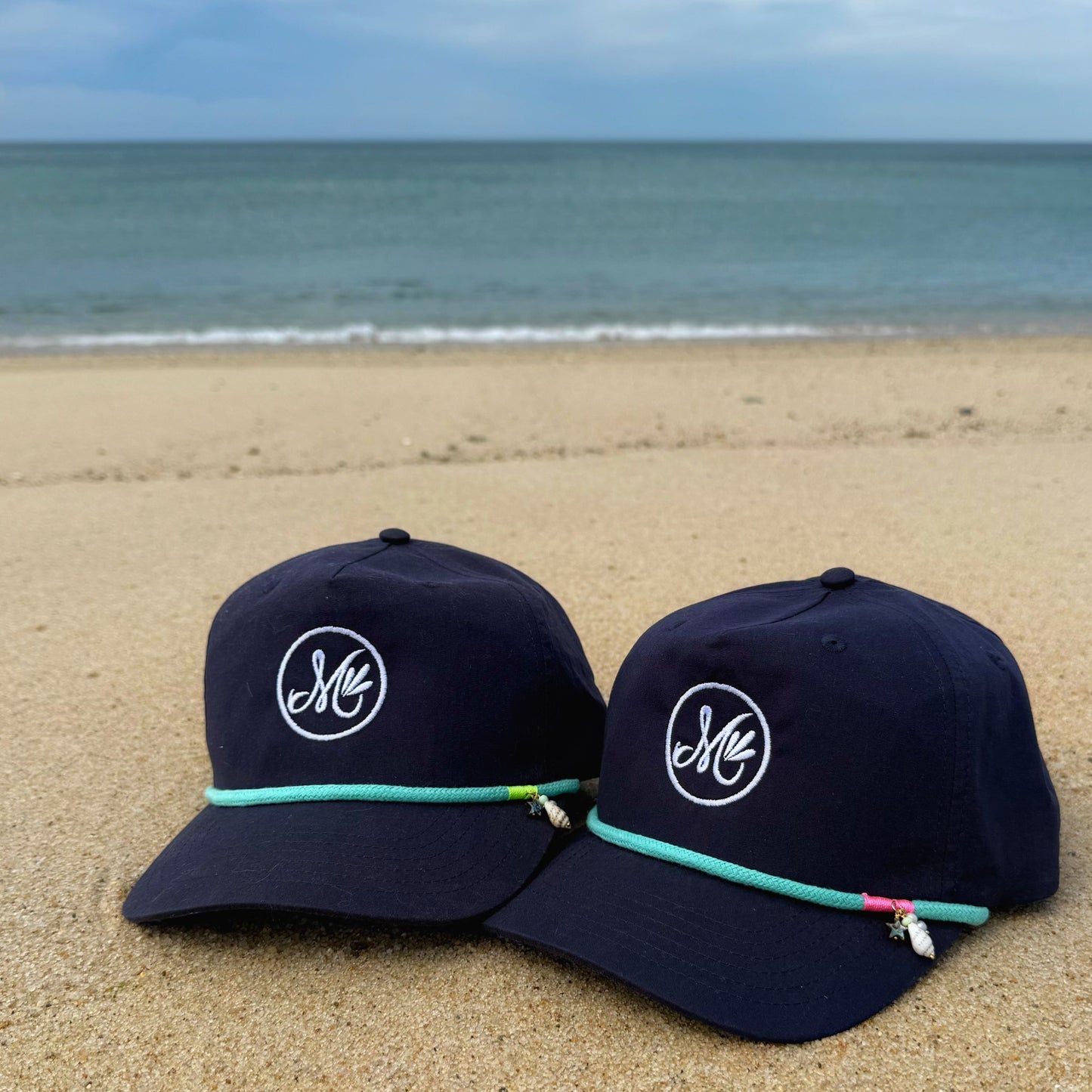 Two navy blue caps with a logo on a sandy beach with ocean in the background