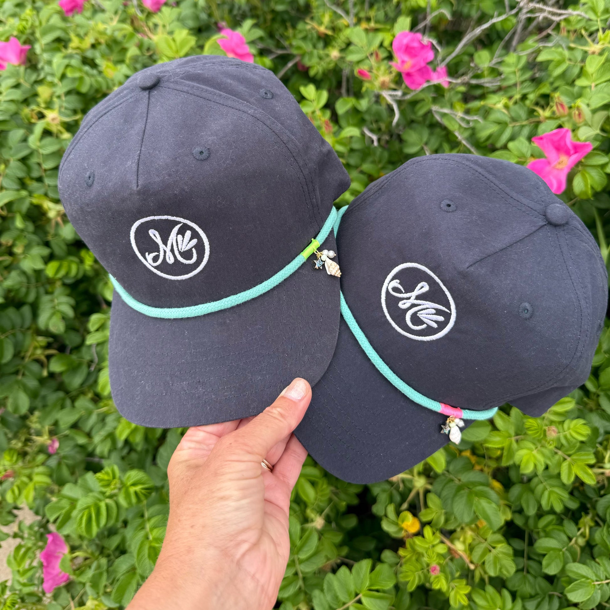 Two black baseball caps with a logo held in front of green foliage and pink flowers.