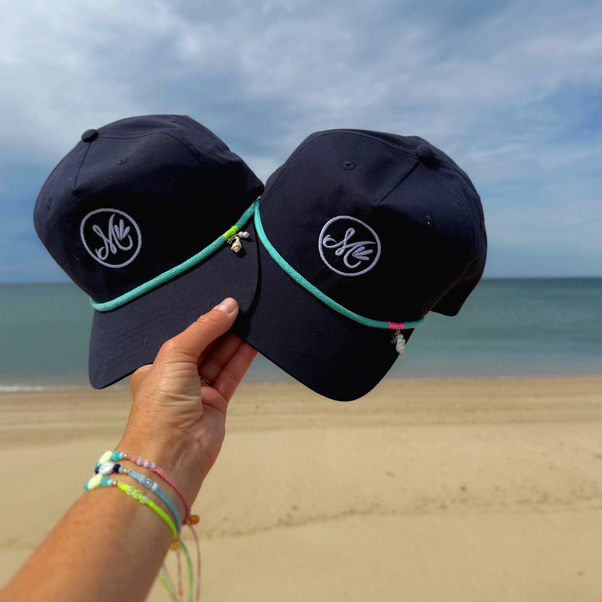 Two navy blue caps with a logo held by a hand on a beach.