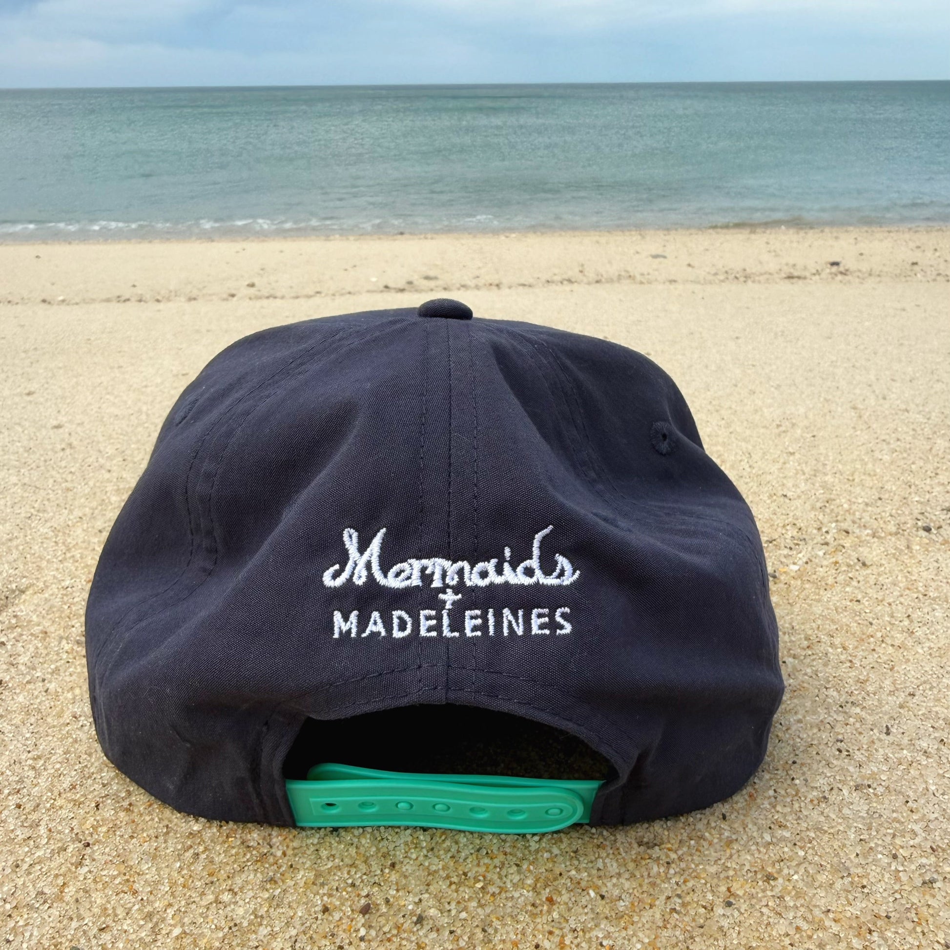 Black cap with 'Mermaids Madeleines' text on a sandy beach with ocean in the background