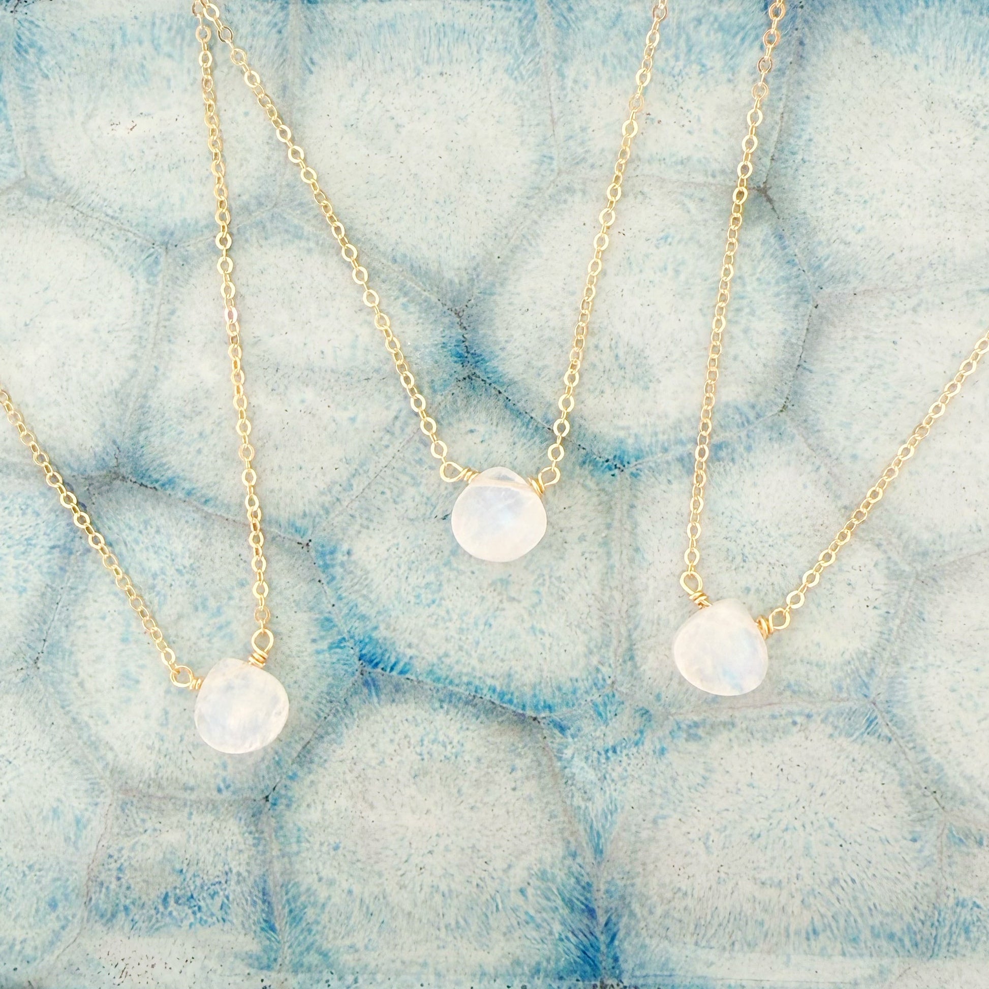 the mermaids and madeleines marshmallow necklace is a dainty 14k gold filled chain necklace with a tiny moonstone drop at the center. this is a photograph of 3 of the necklaces close up on a light blue ceramic dish