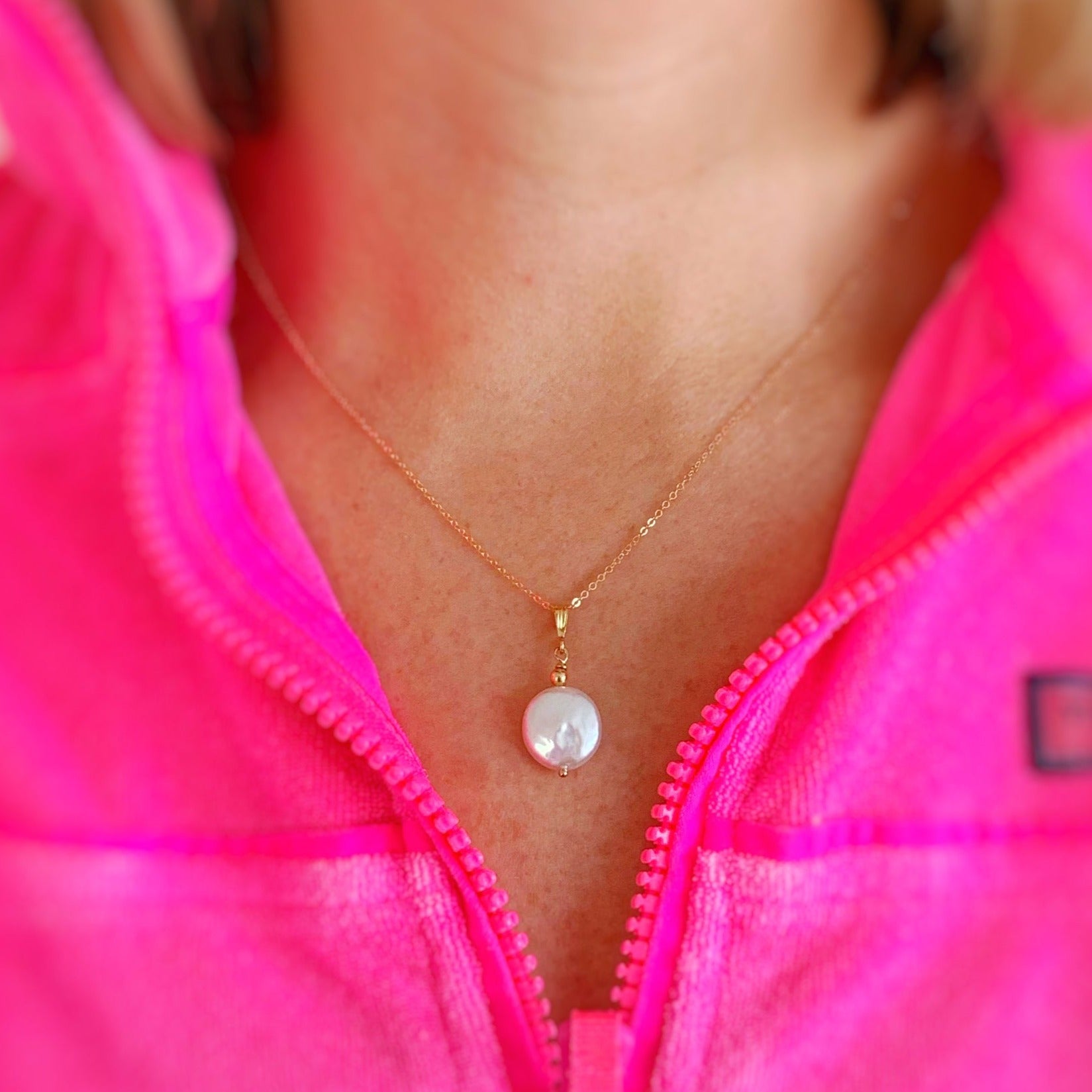 Mermaids and Madeleines Newport Gala necklace in 14k gold filled is created with a large freshwater coin pearl. This pendant style necklace is photographed here on a person with a bright pink quarter zip top