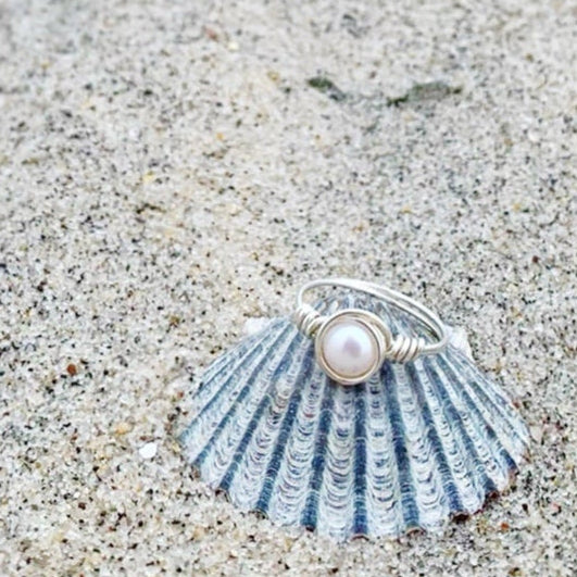 The newport ring by mermaids and madeleines is a sterling silver wire-wrapped style ring that has sterling silver wire swirled around a white freshwater button pearl. this ring is photographed on a scallop shell in the sand.