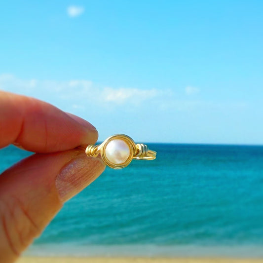 Gold ring with a pearl center held against a beach and blue sky background