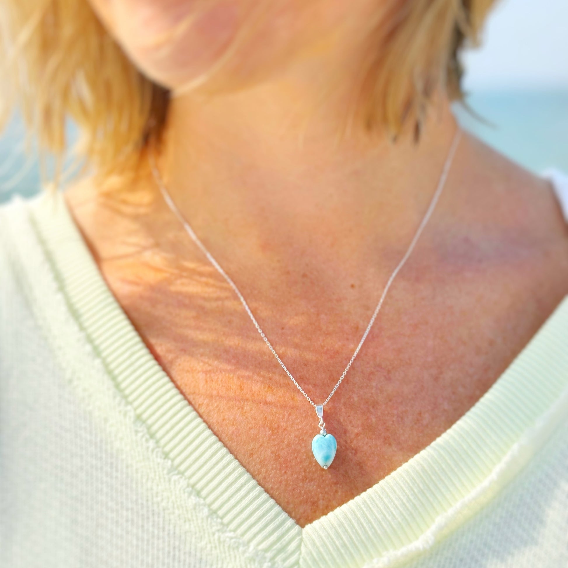 the ocean lover necklace by mermaids and madeleines is a sterling silver pendant style necklace with a heart shaped larimar bead drop. this necklace is photographed on a person close up with a beach background
