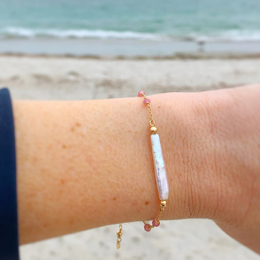 the pink azalea bracelet by mermaids and madeleines is an adjustable chain style bracelet with a slide bead clasp. At the center of the bracelet is a freshwater stick pearl, with pink tourmaline small saucer beads in stations on the 14k gold filled chain. this bracelet is photographed on a wrist with a cloudy beach day in the background