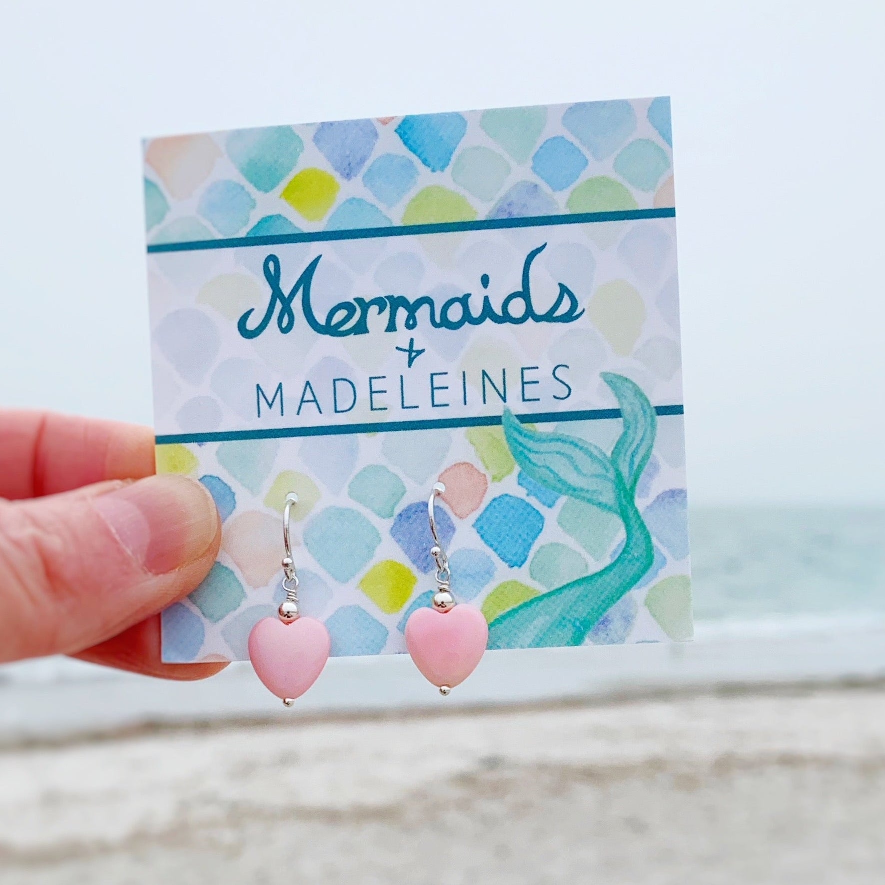 Pink earrings on a 'Mermaids & Madeleines' branded card held by a hand with a beach background.