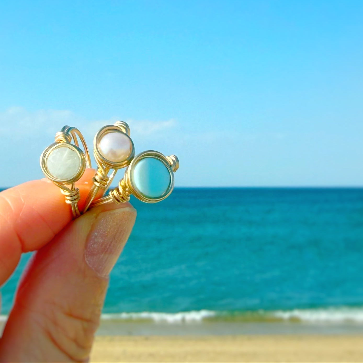 Gold filled wire-wrapped rings with gemstones held against a beach backdrop