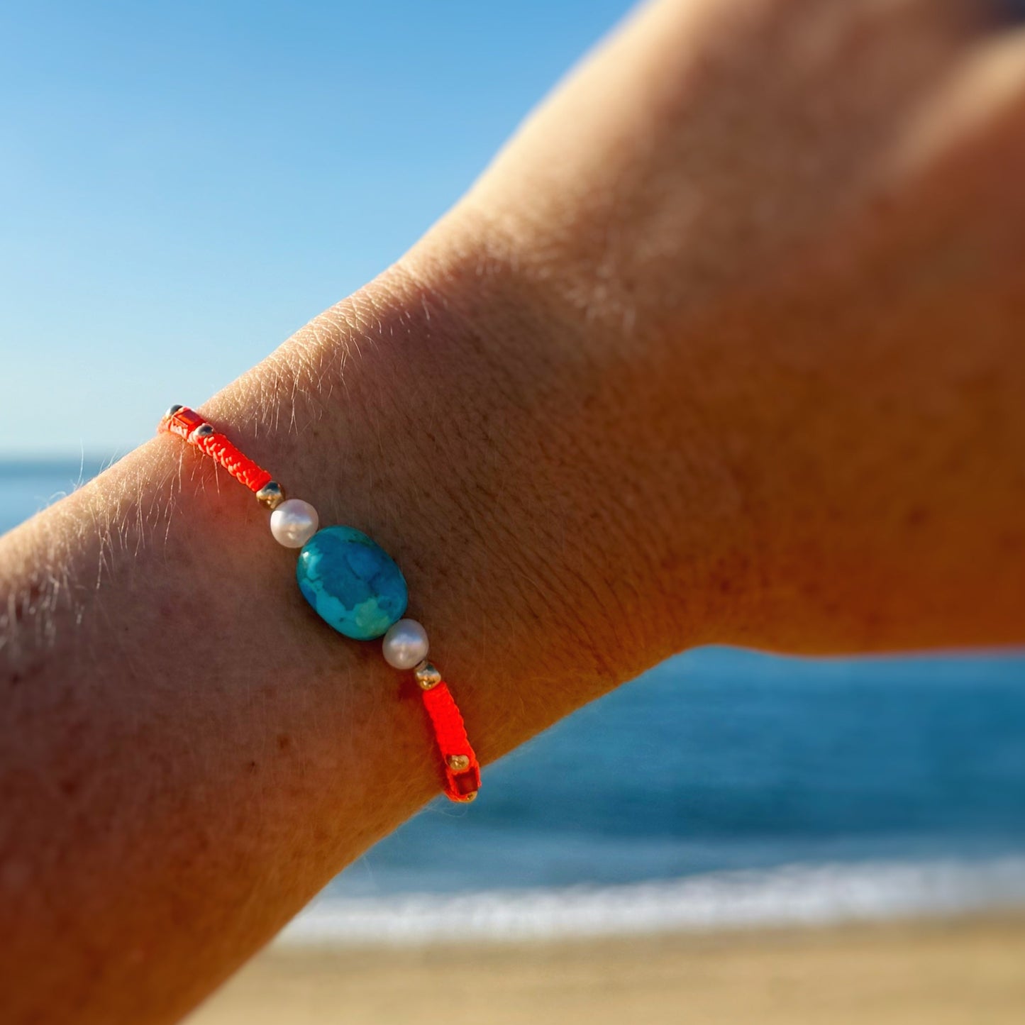 the sedona sunset adjustable macrame bracelet by mermaids and madeleines is a neon orange friendship style bracelet that features a kingman turquoise bead at the center with 2 freshwater pearls, carnelian and 14k gold filled beads and a slider bead clasp. this bracelet is photographed on a wrist with a beach during the daytime in the background