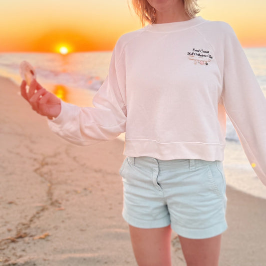 A person standing on a beach at sunset wearing a white sweatshirt with a small navy blue graphic on the left chest and light blue shorts.