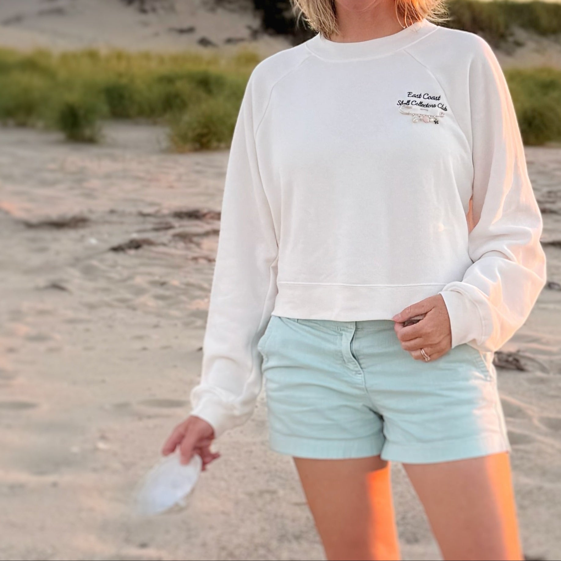 A woman standing on a sandy beach at sunset wearing a white cropped sweatshirt with a small navy embroidered text on the left chest and light mint blue shorts.