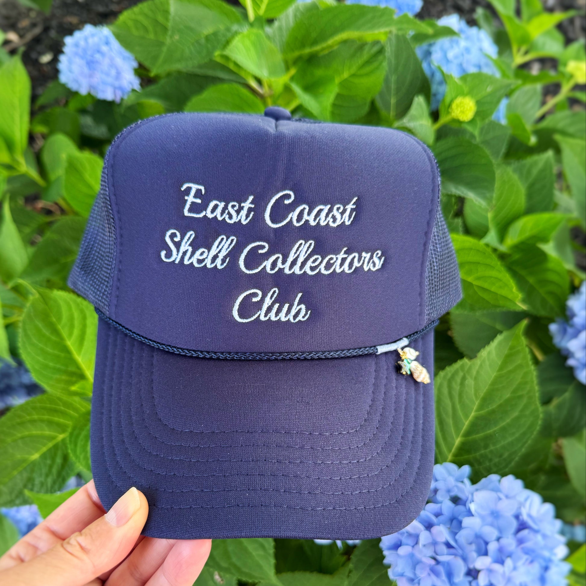 A navy blue trucker hat with 'East Coast Shell Collectors Club' embroidered in white on the front, held by a hand against a background of green hydrangea flowers.