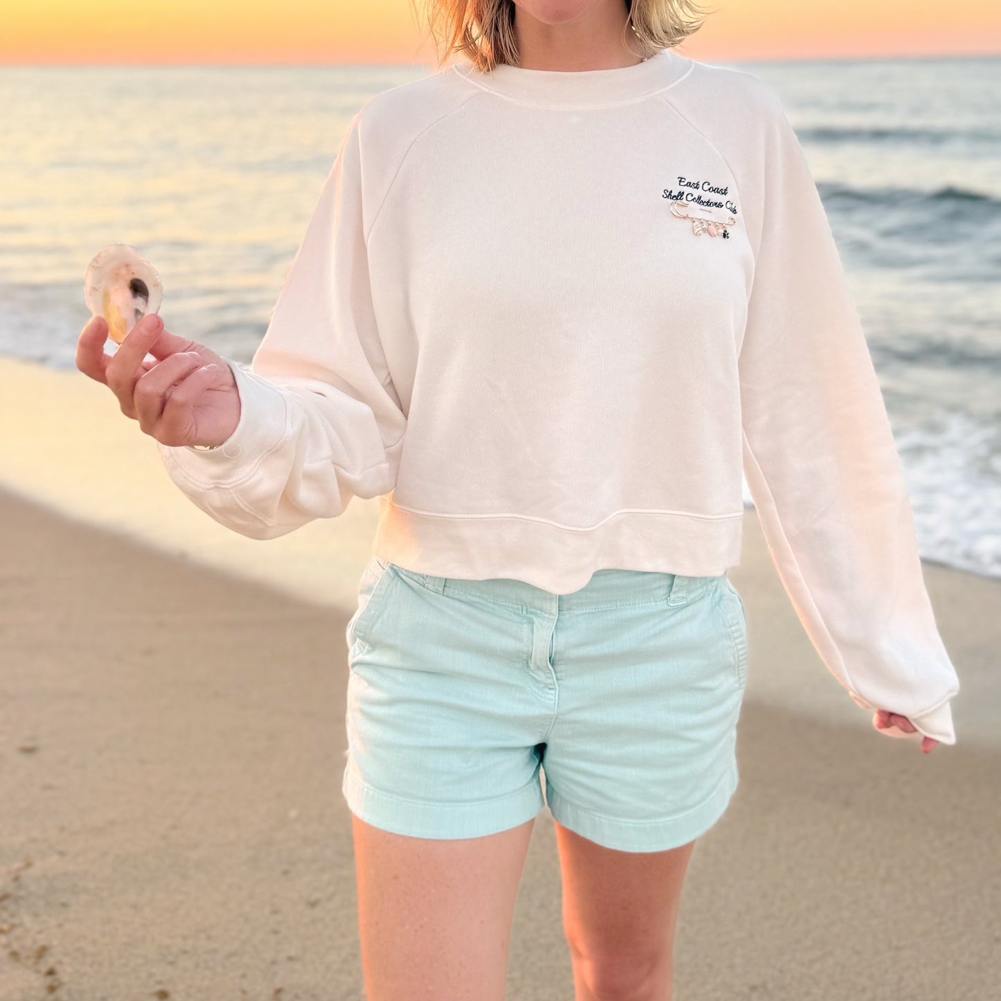 A person standing on a beach at sunset wearing a cream cropped sweatshirt with small navy text graphic on the left chest and light mint shorts, holding a seashell.
