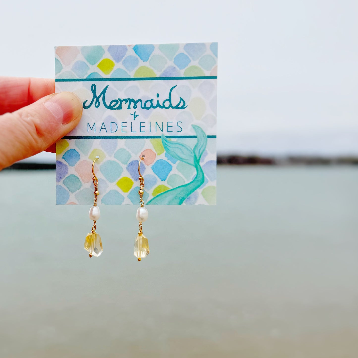 Earrings on a 'Mermaids + Madeleines' card held by a hand against a light background