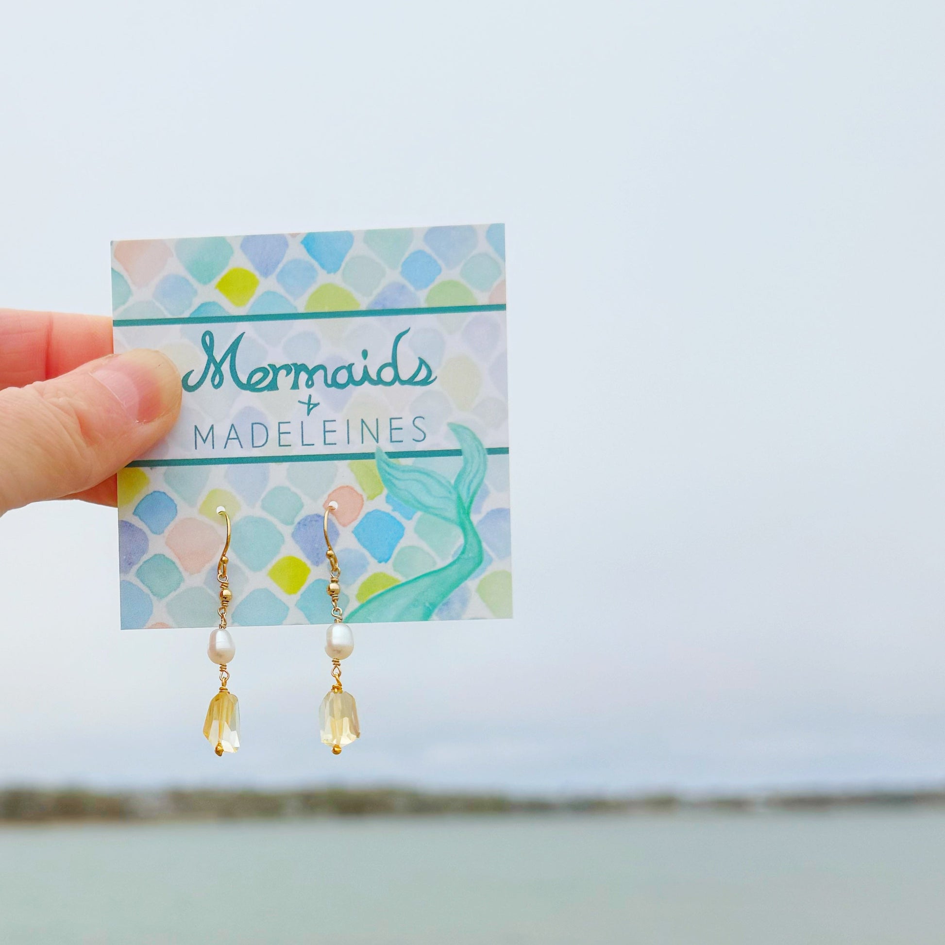 Gold earrings with citrine drop on a 'Mermaids & Madeleines' branded card held by a hand against a light blue ocean background.