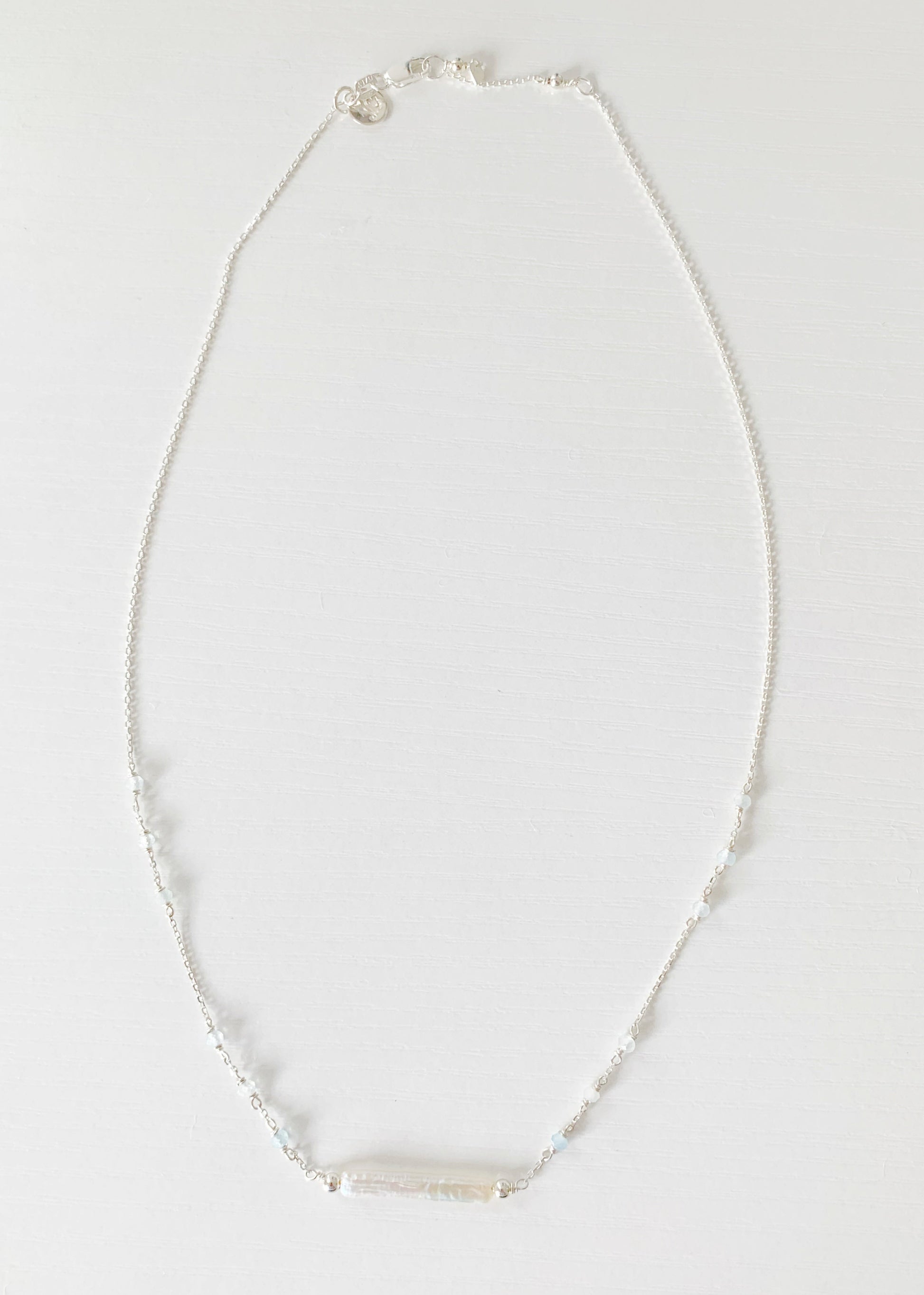 the truro necklace by mermaids and madeleines is a sterling silver chain necklace with a freshwater stick pearl in the center and aquamarine beads going up the sides. this necklace is photographed flat, with a full length view on a white surface