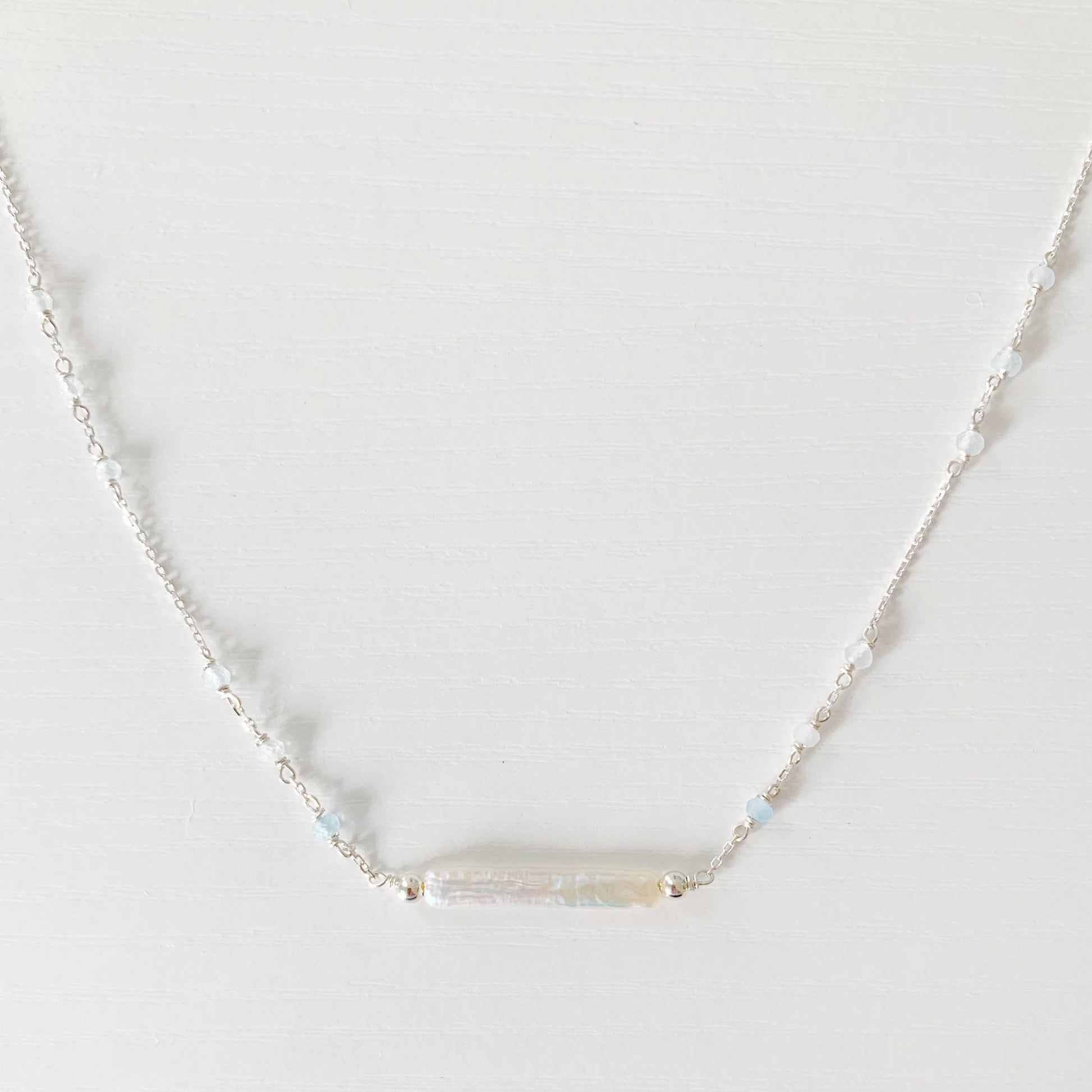 the truro necklace by mermaids and madeleines is a sterling silver chain necklace with a freshwater stick pearl at the center and aquamarine beads going up the sides of the chain. this necklace is photographed close up on a white surface