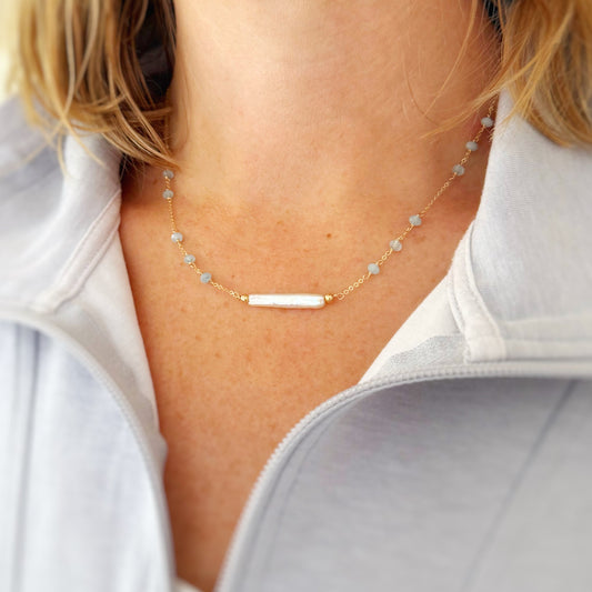 Truro Necklace by mermaids and madeleines is a bar style necklace that has a freshwater stick pearl at the center with small faceted aquamarines on the gold chain. this necklace is photographed on a neck to show how the necklace sits
