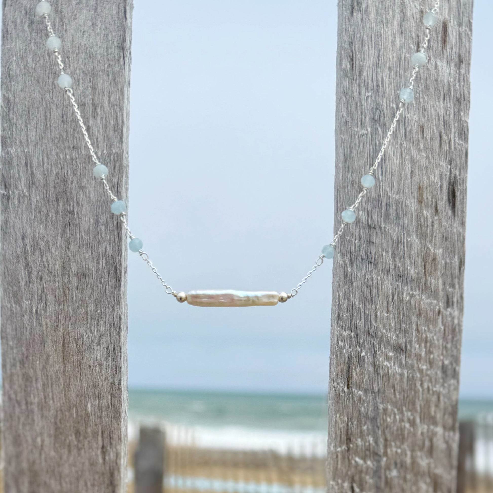 Necklace with a horizontal stick pearl hanging between two wooden posts with a beach in the background