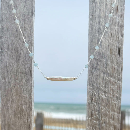 Necklace with a horizontal stick pearl hanging between two wooden posts with a beach in the background