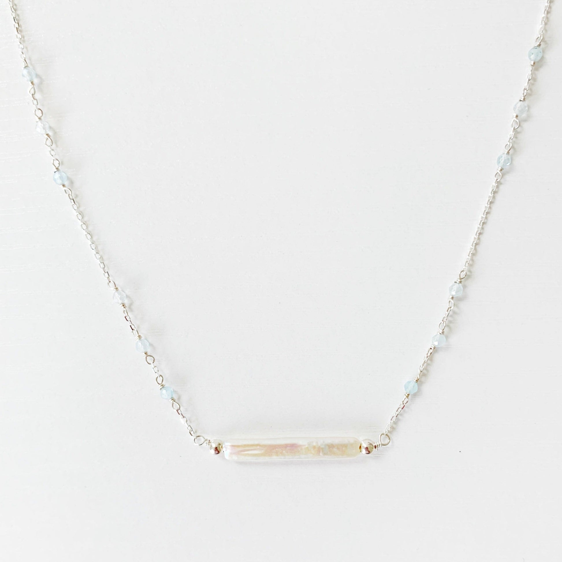 The Truro Necklace by Mermaids and Madeleines features a freshwater stick pearl at the center like a horizontal bar necklace with tiny microfaceted aquamarine beads going up the chain on either side. the chain and findings are sterling silver. this necklace is photographed on a white surface
