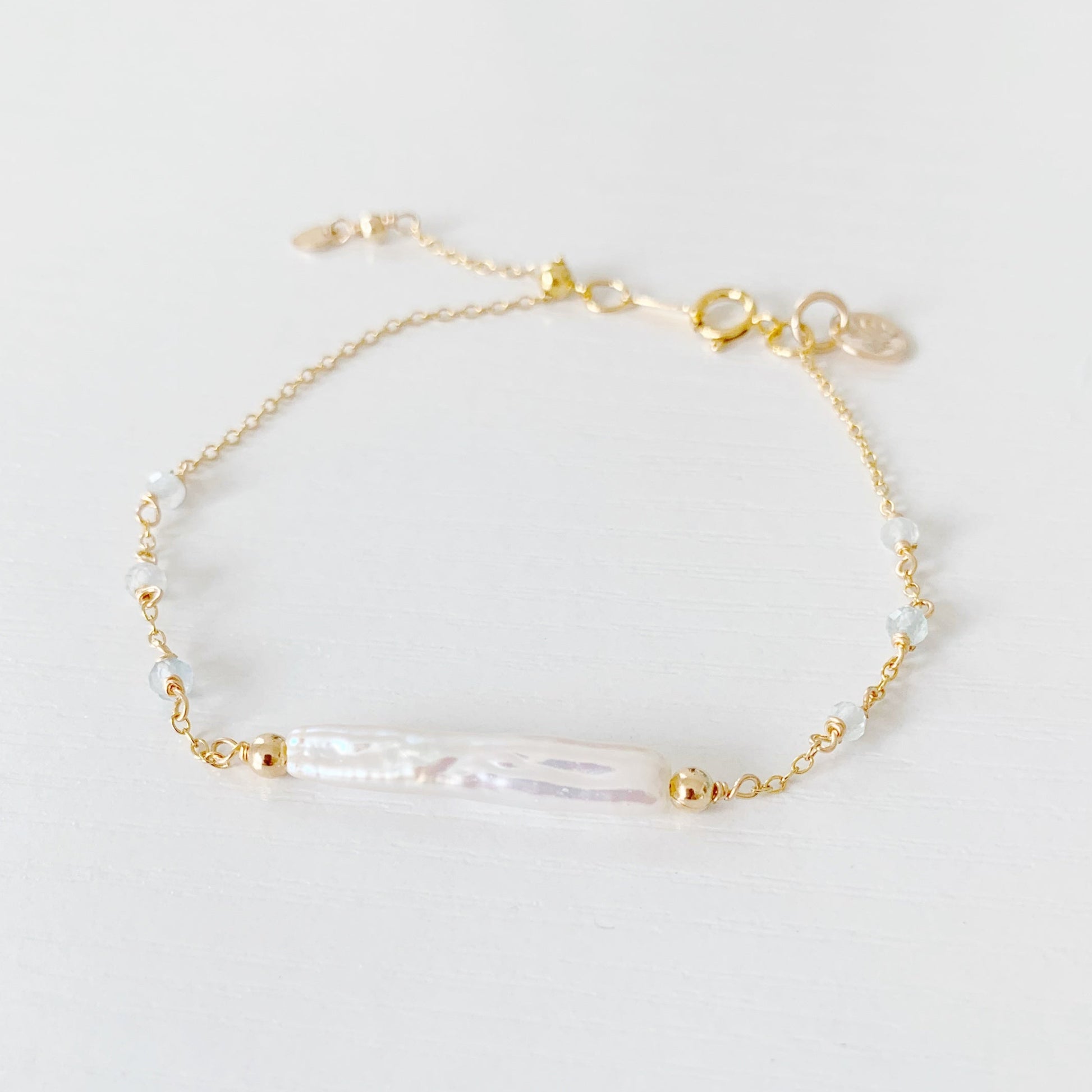 the truro bracelet by mermaids and madeleines is a 14k gold filled chain bracelet with a freshwater stick pearl at the center and aquamarine faceted beads going down the sides. there is a slide bead near the clasp to make the bracelet adjustable. this bracelet is photographed flat on a white surface