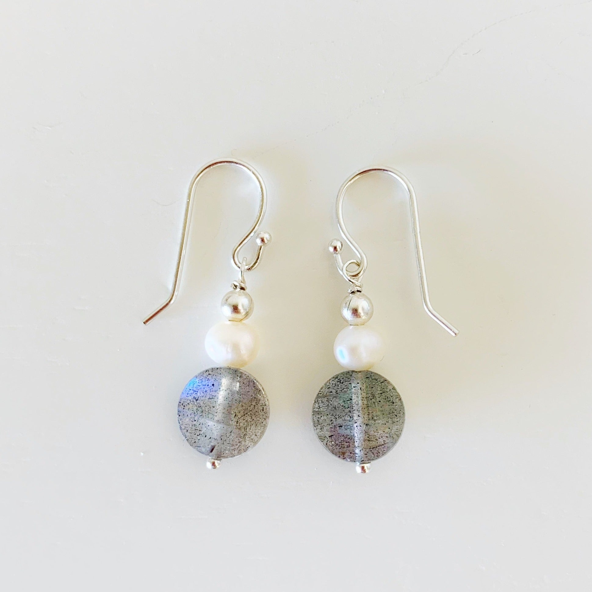 Arctic earrings by mermaids and madeleines are created with freshwater pearls, and faceted labradorite coins and sterling silver findings. this pair is photographed on a white surface