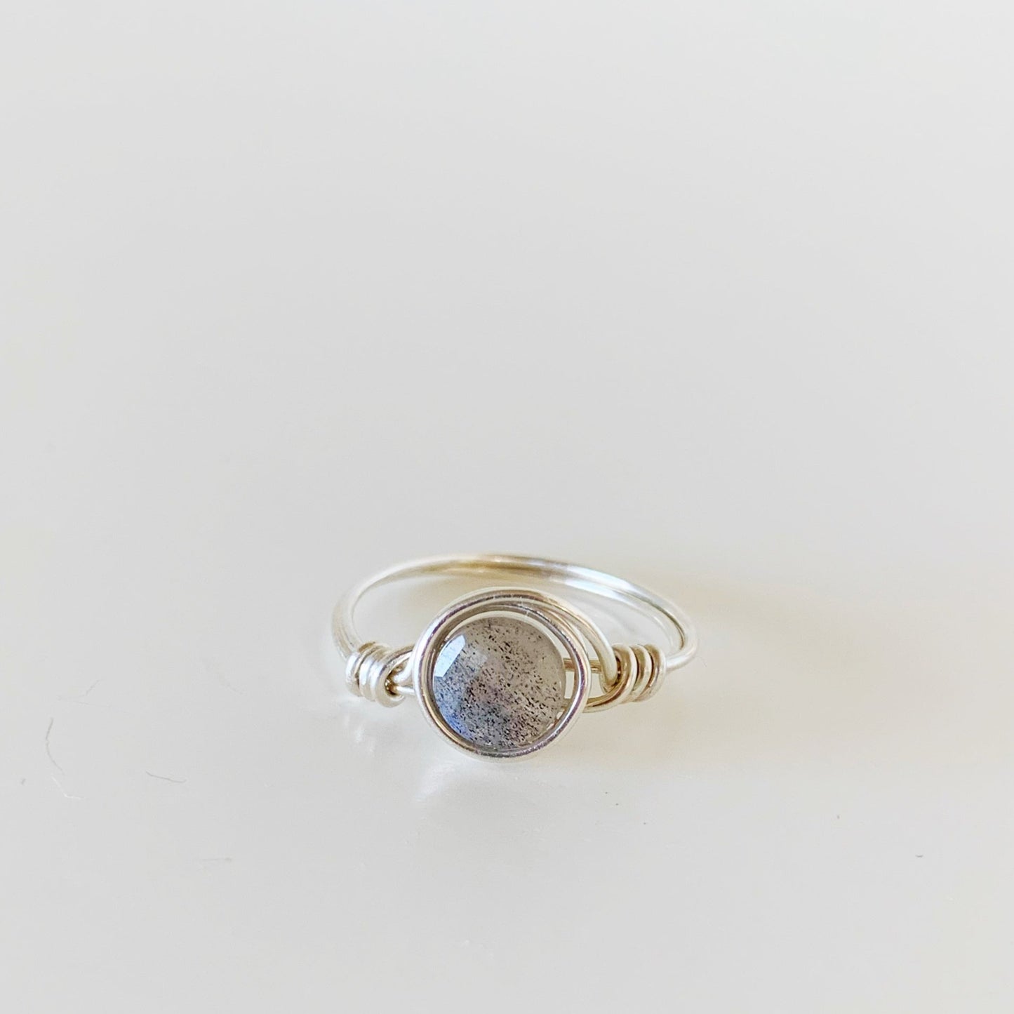 the mermaids and madeleines arctic ring is a wire-wrapped ring created with sterling silver wire and a faceted coin labadorite bead at the center. this ring is facing forward and photographed on a white surface