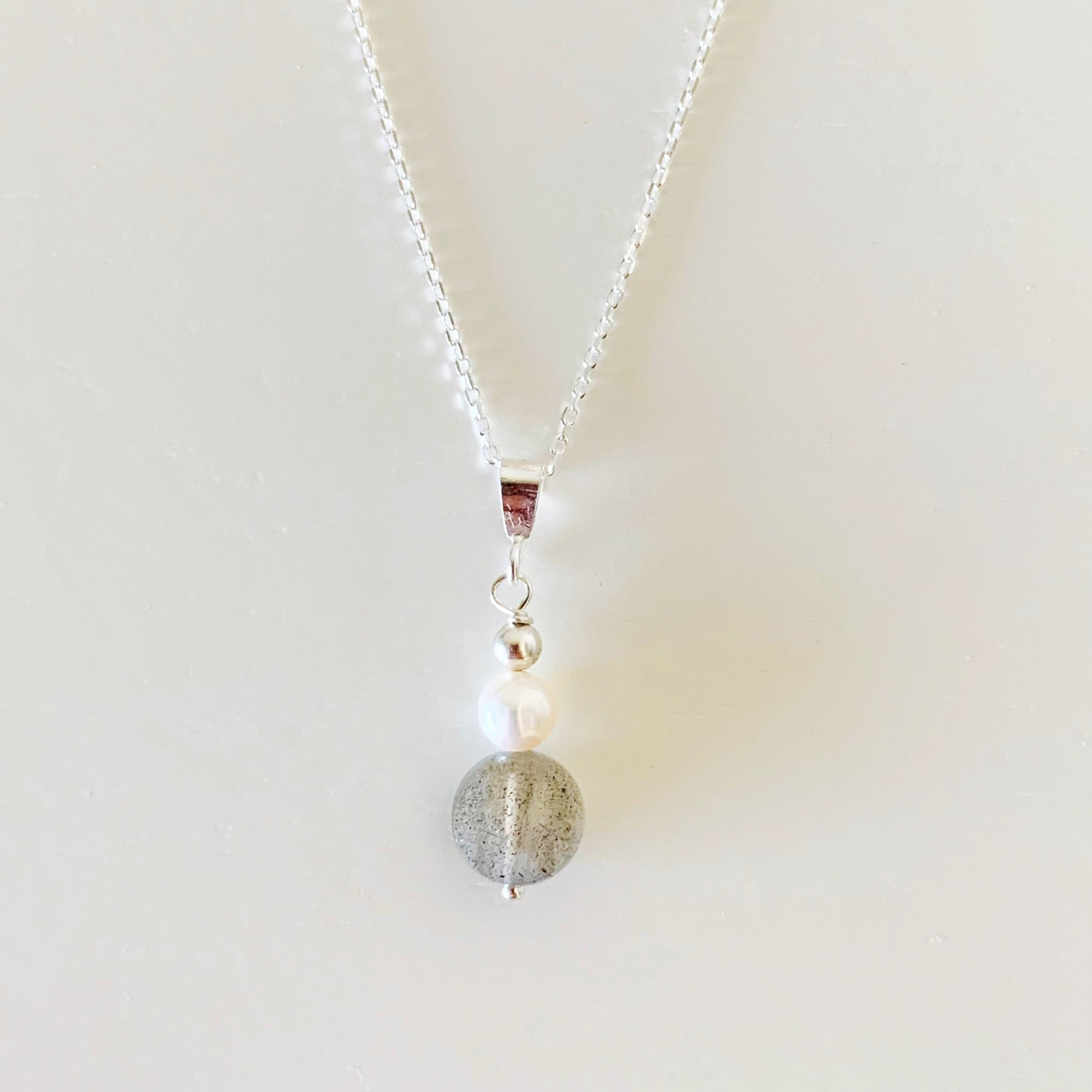 The arctic pendant necklace by mermaids and madeleines is created with a freshwater pearl and a faceted labradorite coin bead and features sterling silver findings and chain. this piece is photographed on a white surface