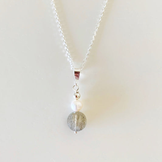 The arctic pendant necklace by mermaids and madeleines is created with a freshwater pearl and a faceted labradorite coin bead and features sterling silver findings and chain. this piece is photographed on a white surface