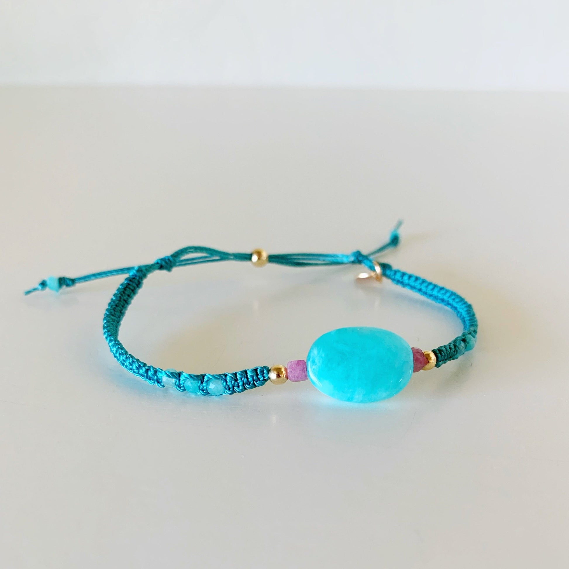 the harwich port adjustable macrame bracelet by mermaids and madeleines in color blue spruce is a friendship style bracelet with a slide bead clasp and a bright amazonite bead at the center with small amazonite rondelles on the side of the bracelet. this piece is complimented by pink tourmaline and 14k gold filled beads. this bracelet is facing to the right and photographed on a white surface