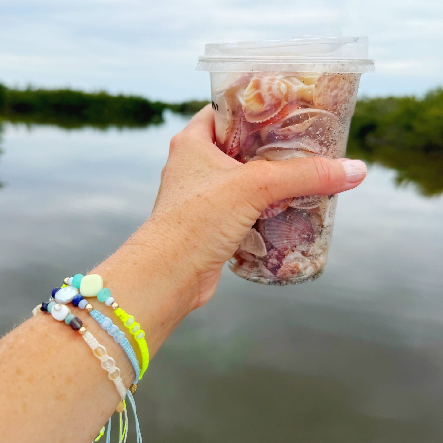 Hand holding a cup with seashells against a blurred natural background. Wrist has 3 woven bracelets on it.