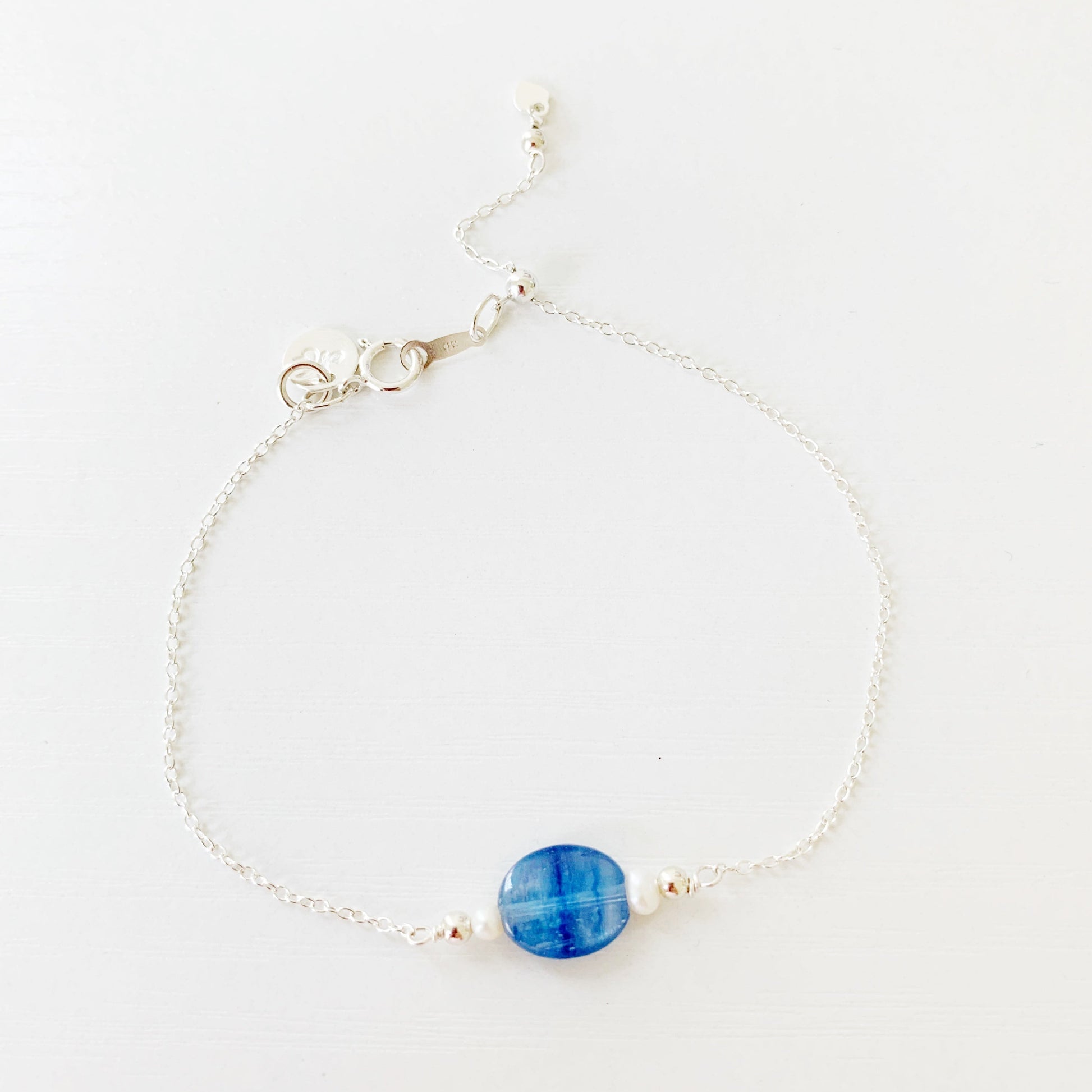 the brewster bracelet by mermaids and madeleines is an adjustable sterling silver chain bracelet with a slide bead at the clasp. there's a blue kyanite coin bead at the center complimented by small freshwater pearls on each side. this bracelet is photographed from the top down view on a white surface