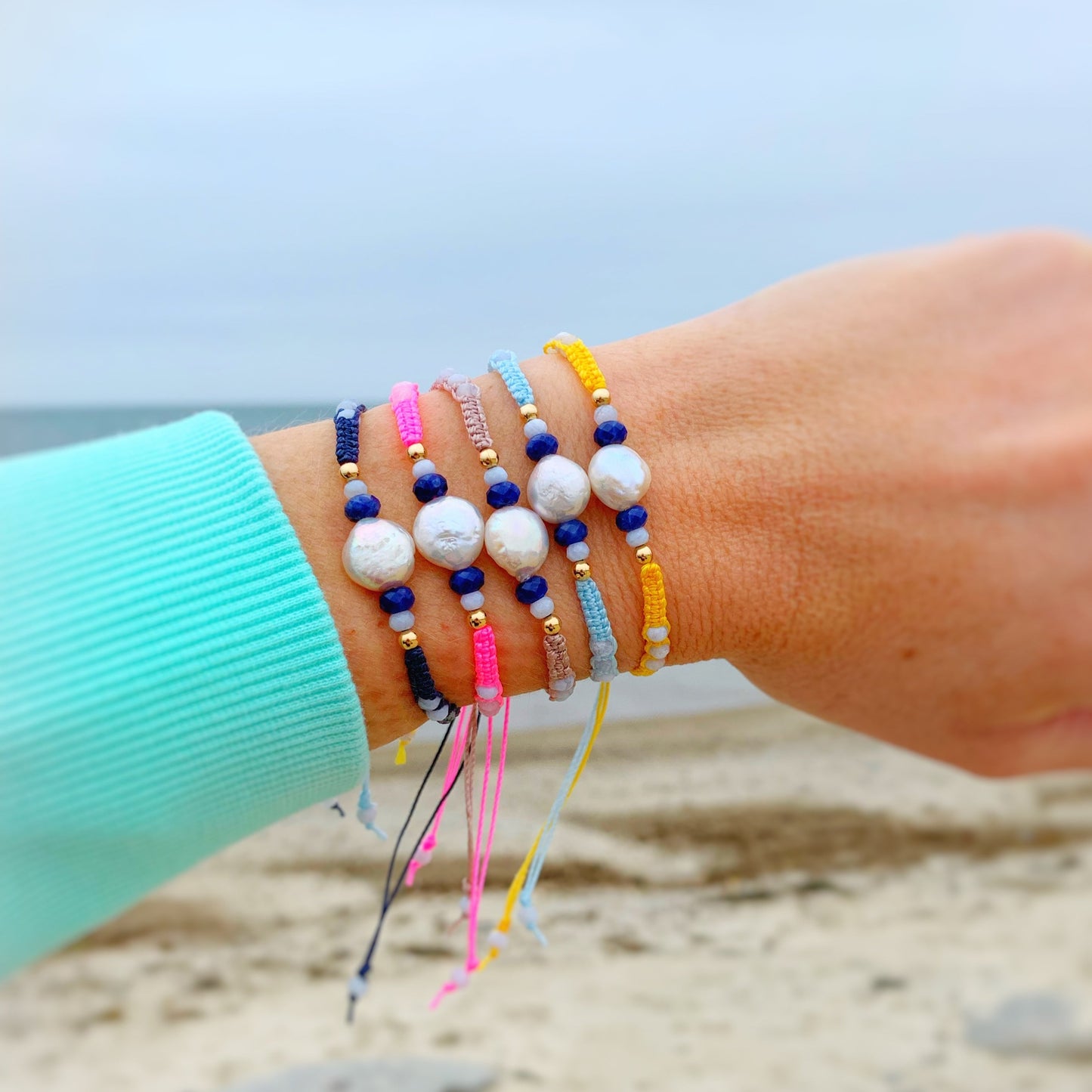 Mermaids and madeleines bristol macrame bracelets all four color stacked up and worn on a wrist in front of the beach in the background