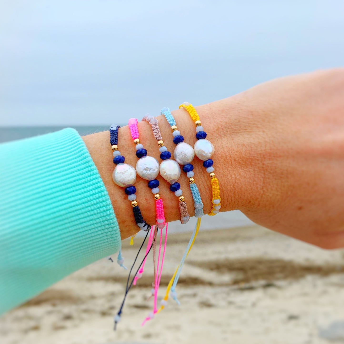 Bristol macrame bracelets by mermaids and madeleines have a freshwater pearl and semiprecious beads at the center and a 14k gold filled slide bead closure. Here is a photograph of all four bristol bracelets worn stacked on a wrist with the beach and ocean in the background