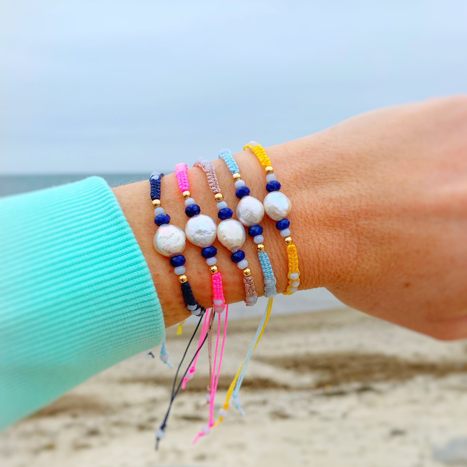 Mermaids and madeleines bristol macrame bracelets are adjustable woven friendship bracelets with a 14k gold filled slide clasp. pictured here are all 5 available colors worn on a wrist in front of the ocean and beach in the background