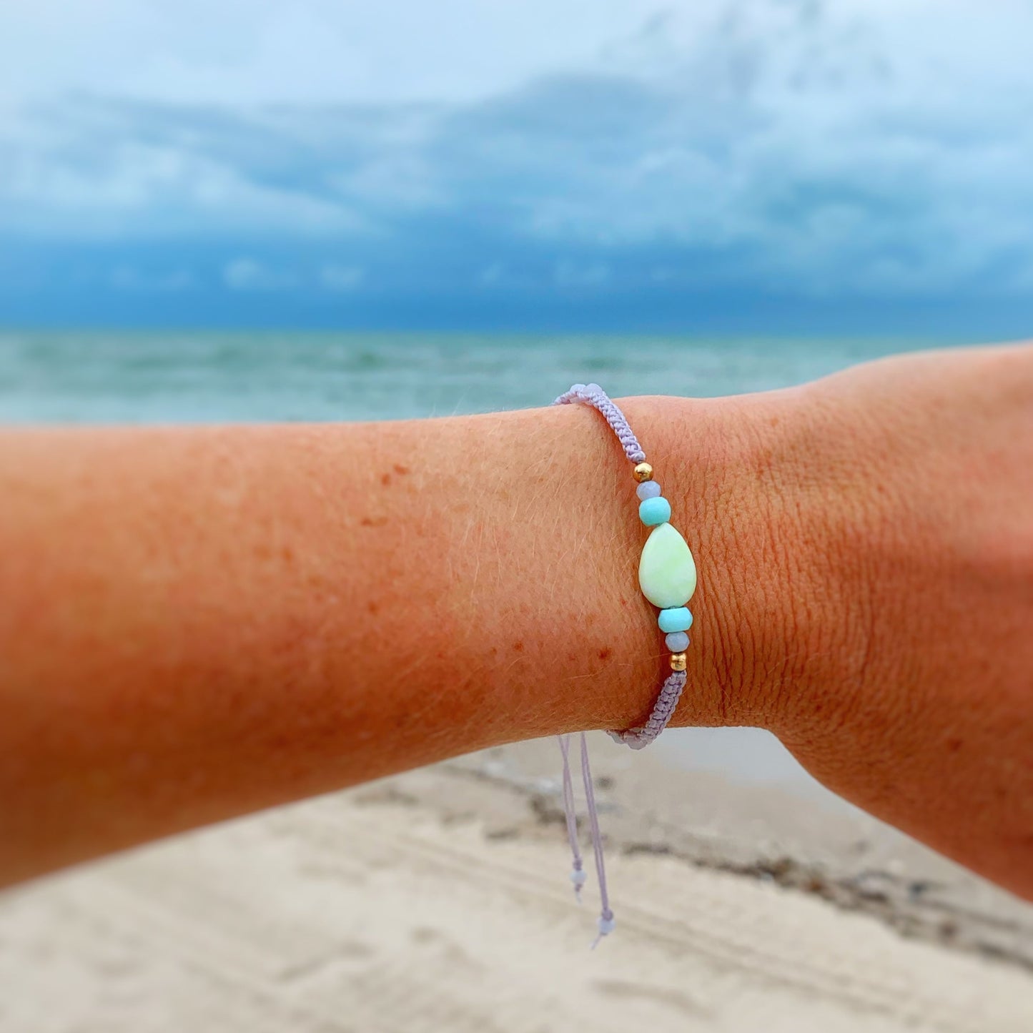 the captiva bracelet in coastal haze by mermaidsand madeleines is a macrame bracelet created with a lime jade teardrop bead at the center with semiprecious and 14k gold beads on either side with lavender color cord. this bracelet is photographed on a wrist with beach scenery in the background