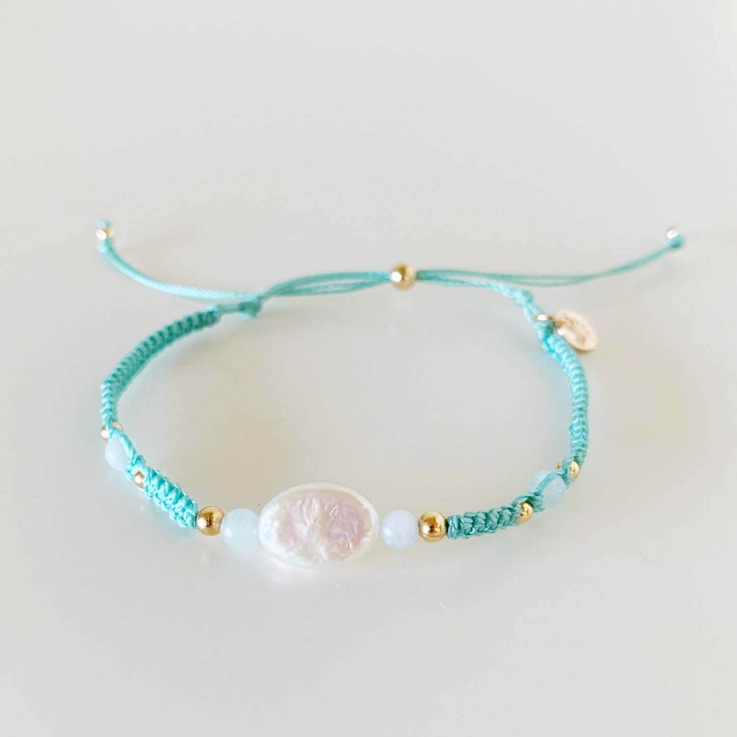 certain urchin is an adjustable macrame bracelet by mermaids and madeleines. its knotted with an icy sage green cord and has an oval shape freshwater pearl at center with 4mm round aqumarine beads on either side. this bracelet is photographed from the front on a white surface