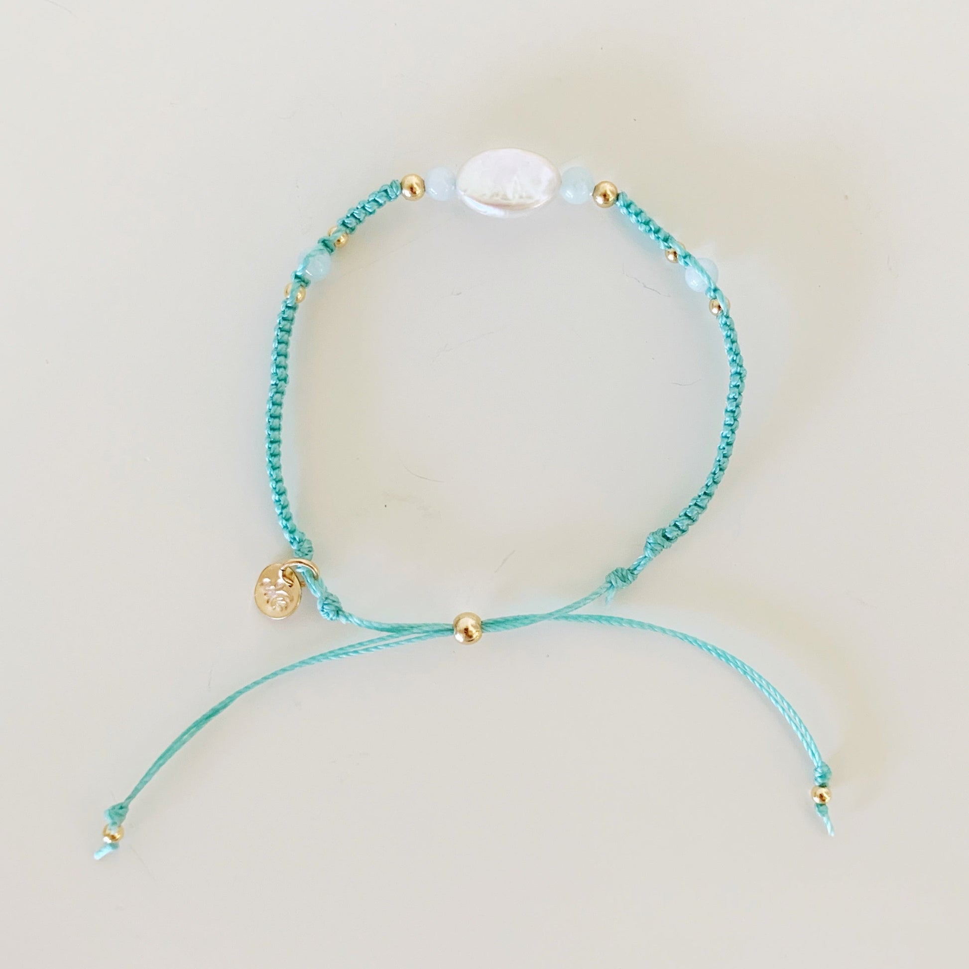 the certain urchin adjustable bracelet by mermaids and madeleines is created with an icy sage green color cord and knotted in a macrame style with a freshwater oval pearl at the center and 4mm aquamarine beads on either side. this bracelet is photographed in a top down full view on a white surface