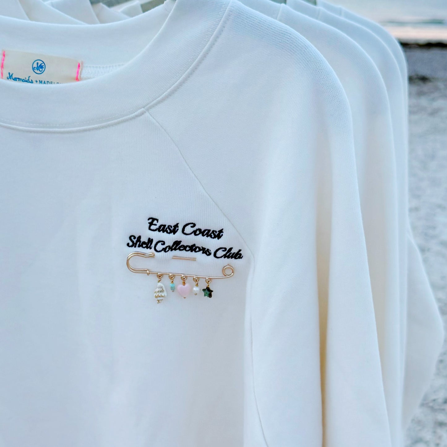 A close-up of a white crewneck sweatshirt hanging on a rack, featuring navy blue embroidered text reading 'East Coast Shell Collectors Club' above a small gold safety pin charm with pastel pink, green, and white beads.