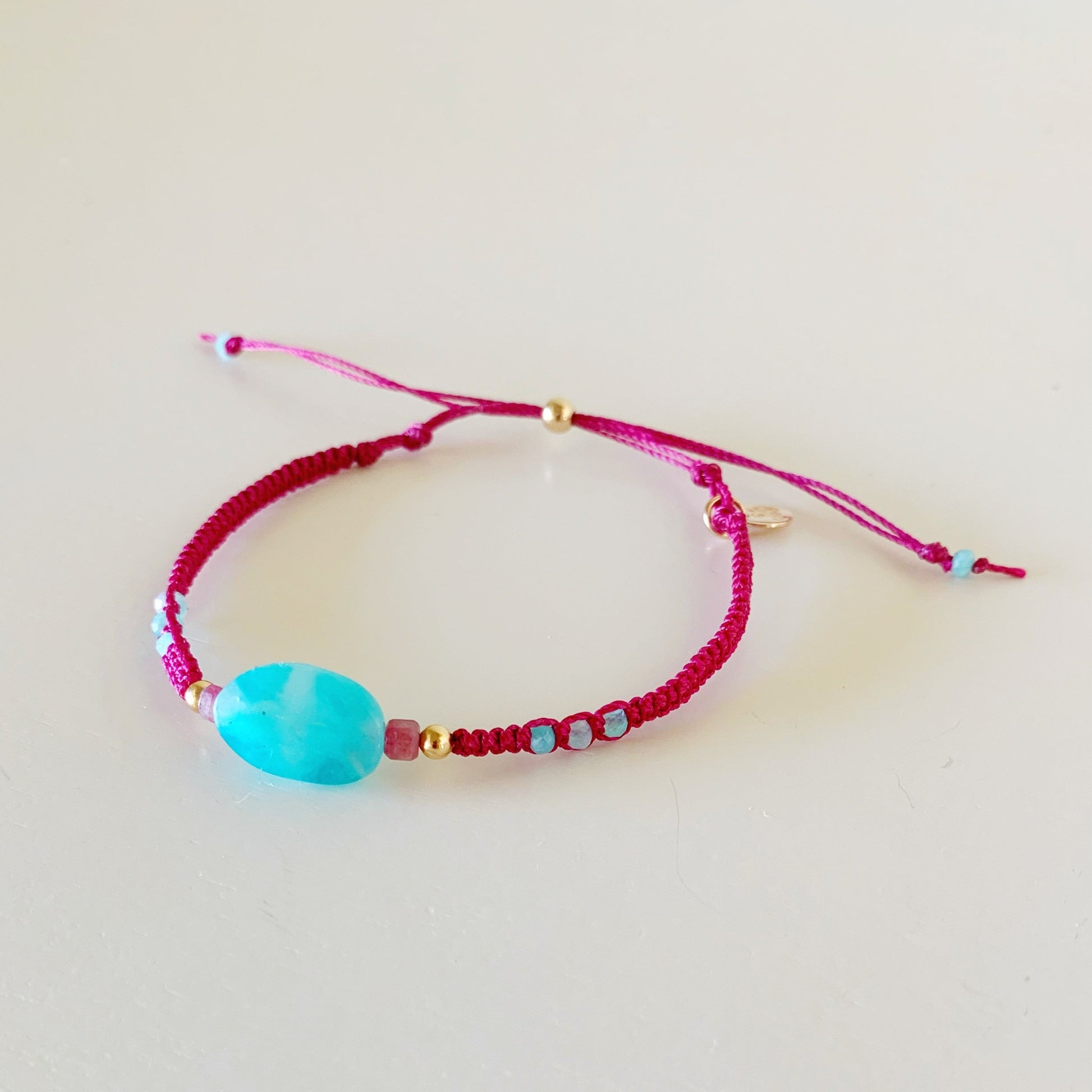 the coastal cranberry harwich port adjustable macrame bracelet by mermaids and madeleines is an adjustable friendship bracelet style with an oval amazonite bead at the center and sides complimented by pink tourmaline and 14k gold filled beads. this bracelet is facing to the left and photographed flat on a white surface