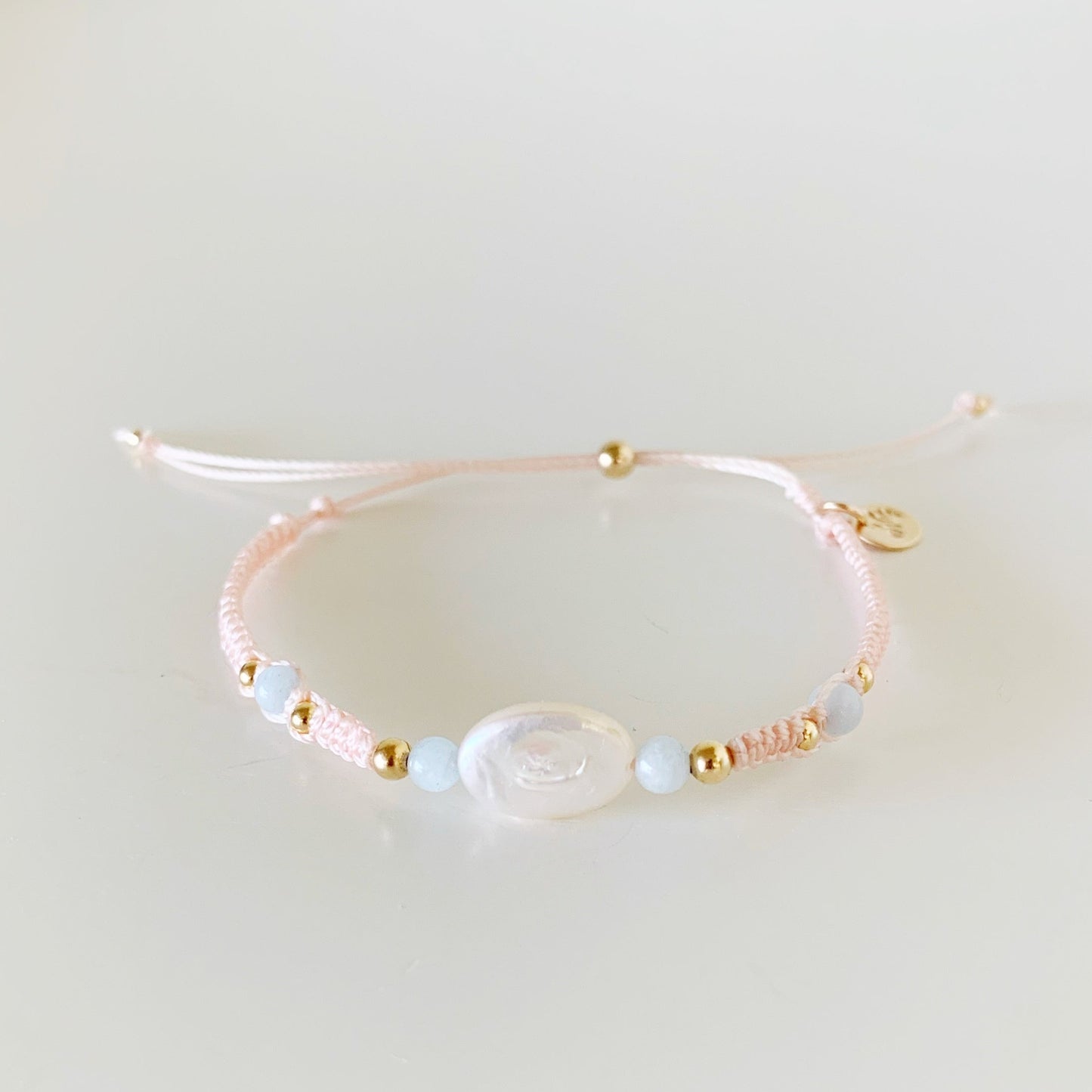 the happy as a clam bracelet by mermaids and madeleines is an adjustable macrame style bracelet designed with a pastel blush color cord with a freshwater oval pearl at the center with 4mm aquamarine beads on eiither side. this bracelet is photographed from the front on a white surface