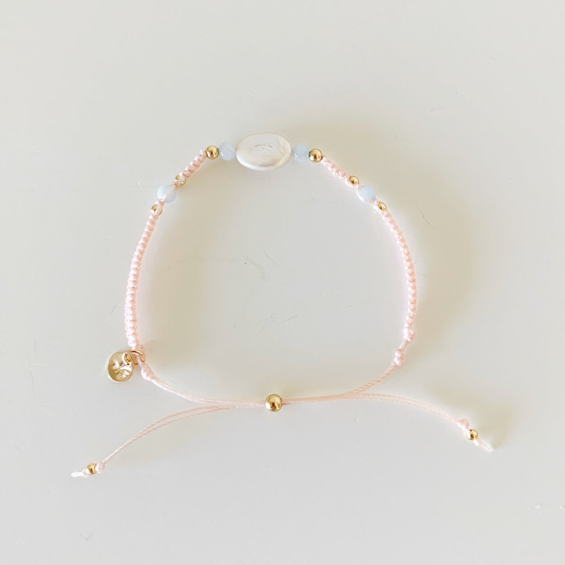 the happy as a clam bracelet by mermaids and madeleines is an adjustable macrame style bracelet created with light pastel blush cord with a freshwater oval pearl at the center with 4mm round aquamarine on either side. this bracelet is photographed from the top in full view on a white surface