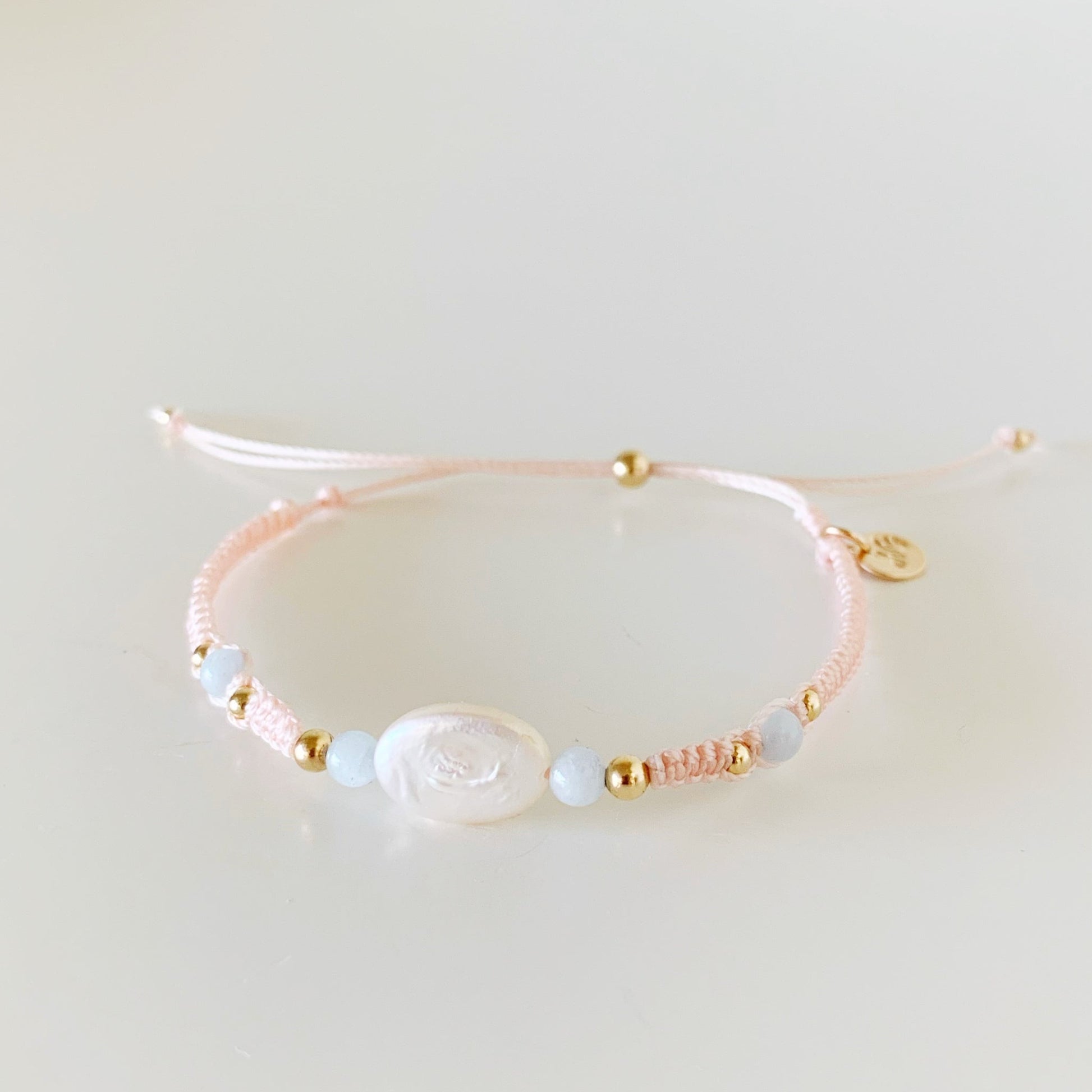 happy as a clam bracelet by mermaids and madeleines is an adjustable macrame style with pastel blush cord with a freshwater oval pearl at the center with 4mm round aqumarine beads on either side. this bracelet is photographed from the front on a white surface
