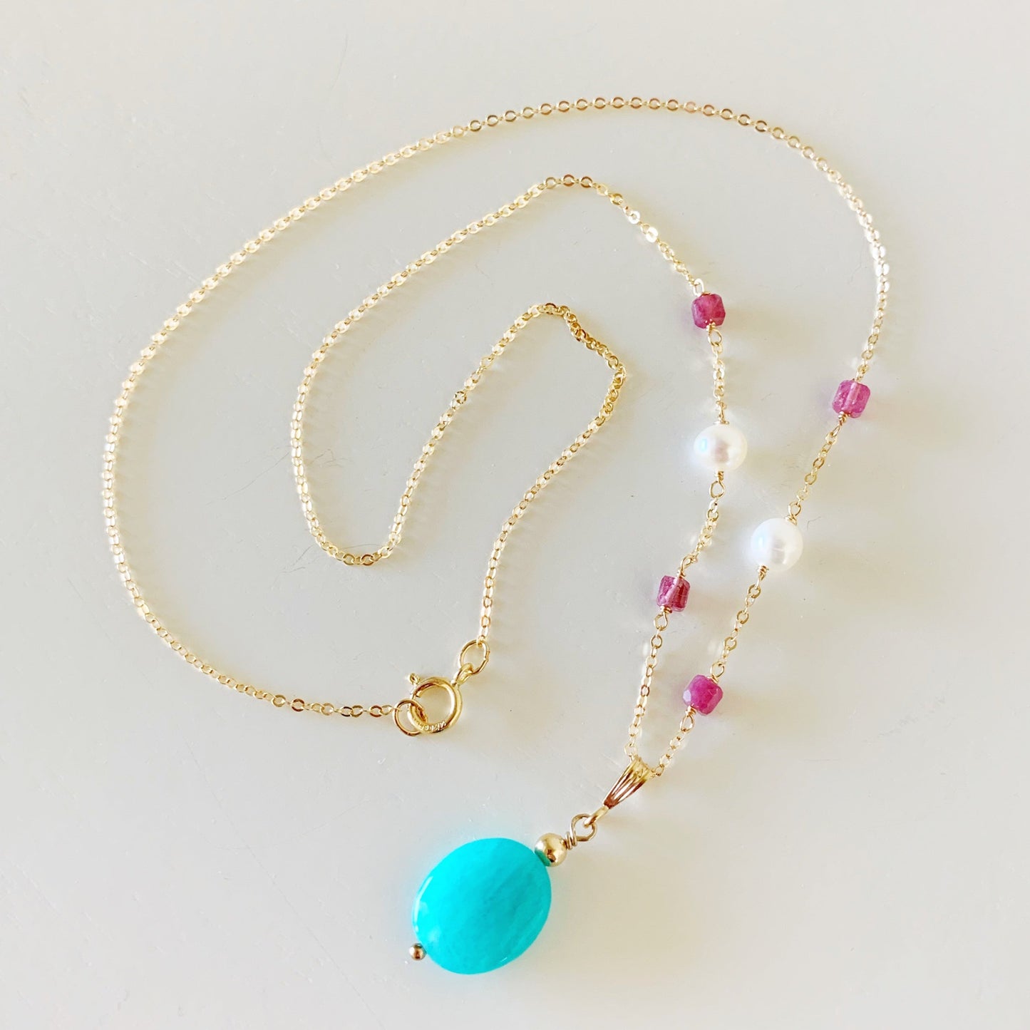 the harwich port necklace by mermaids and madeleines is a pendant style necklace with an amazonite stone suspended from 14k gold filled chain. the chain has pink tourmaline and freshwater pearls wired in. this necklace is coiled and photographed on a white surface