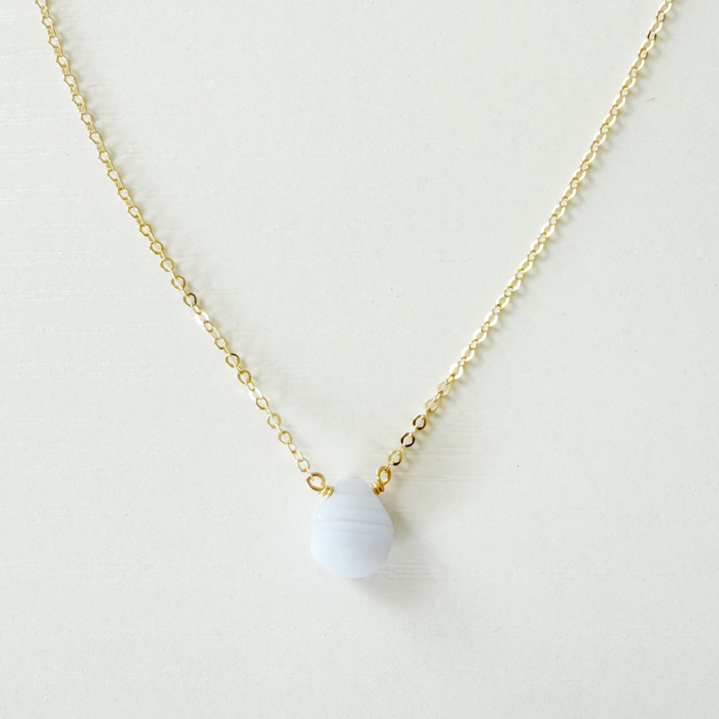A delicate gold chain necklace with a small, smooth, pale blue-gray bead pendant, displayed against a clean, off-white background.