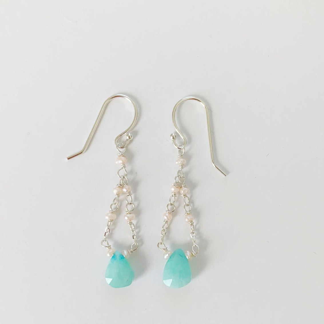 the island air earrings by mermaids and madeleines are created with amazonite teardrop stones at the bottom of the earring with sterling silver chain mixed with pearls looping back up to the ear hook. this pair is photographed on a white background