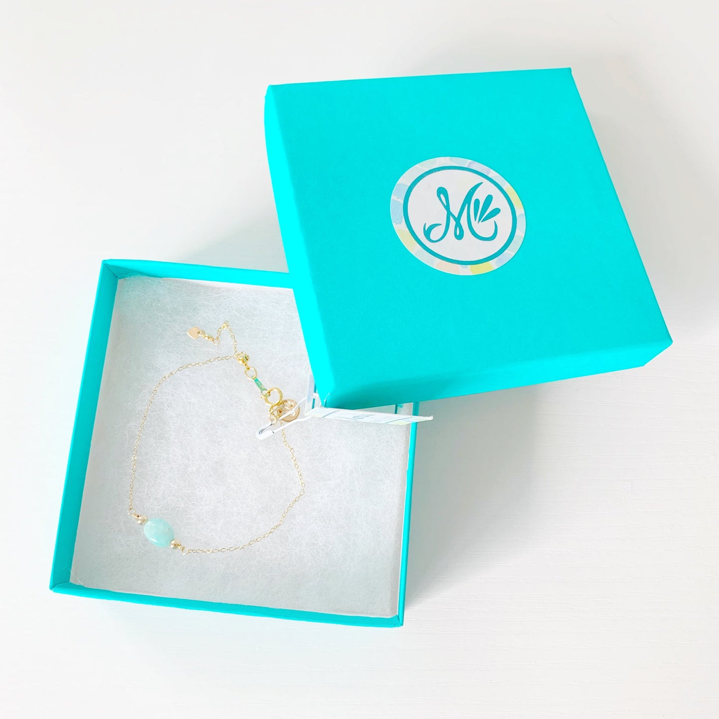 the laguna bracelet by mermaids and madeleines is created with 14k gold filled chain with an aqua, oval amazonite bead at the center and a slide bead at the clasp to make the bracelet adjustable. this bracelet is photographed in a teal gift box on a white table