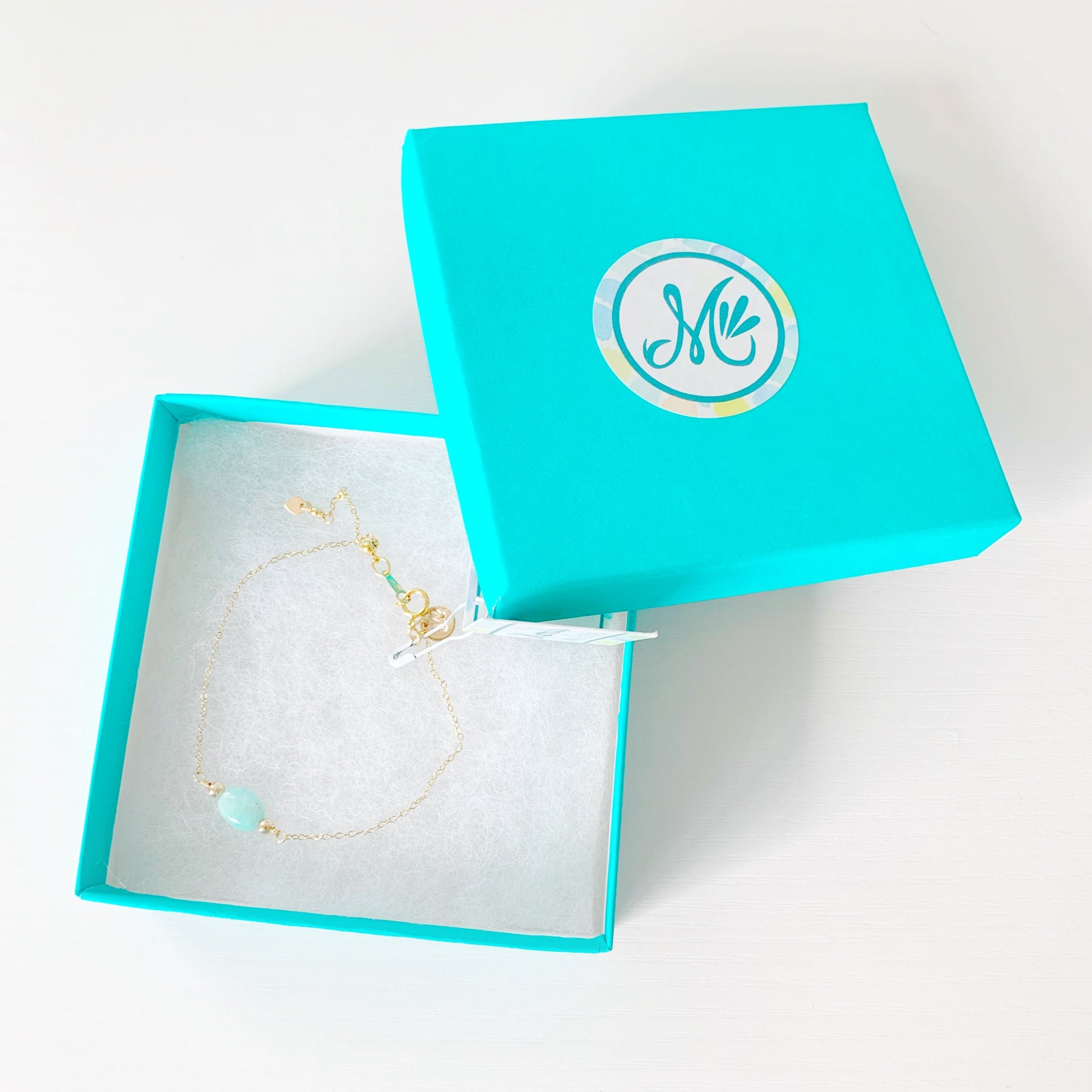 the laguna bracelet by mermaids and madeleines is created with 14k gold filled chain with an aqua, oval amazonite bead at the center and a slide bead at the clasp to make the bracelet adjustable. this bracelet is photographed in a teal gift box on a white table