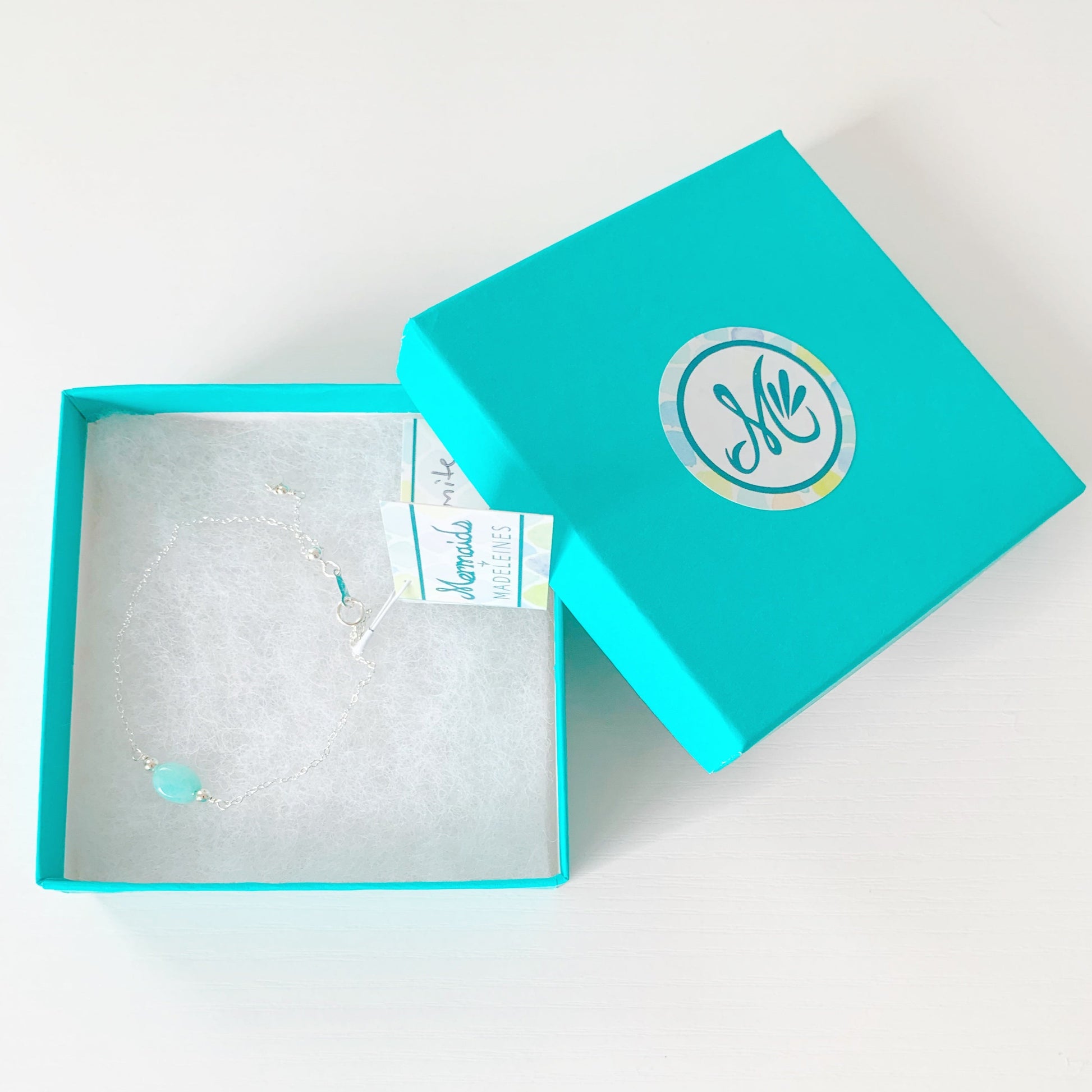 the laguna adjustable bracelet by mermaids and madeleines is created with sterling silver chain with an aqua oval amazonite bead at the center. There's a slide bead at the clasp to make the chain bracelet adjustable. this bracelet is photographed in a teal gift box on a white surface